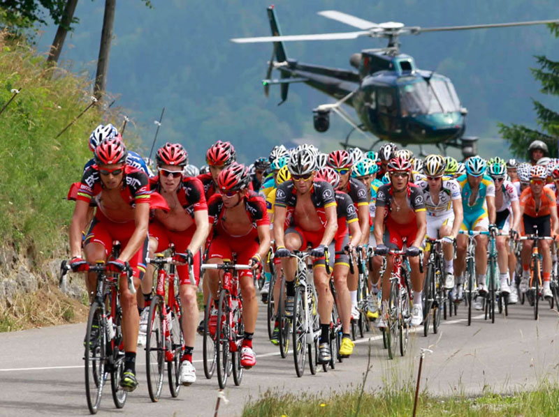 Los ciclistas del Tour de Francia avanzan con dificultad a lo largo de una carretera empinada, un helicóptero desciende en picado detrás para filmarlos.