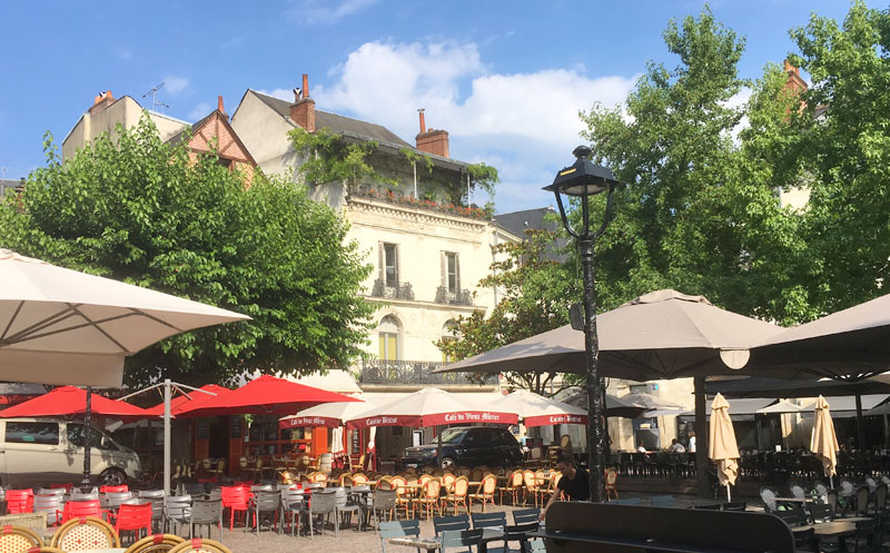 Plaza iluminada por el sol bordeada de árboles y llena de mesas y sillas con sombrillas luminosas, ciudad de Tours, Valle del Loira