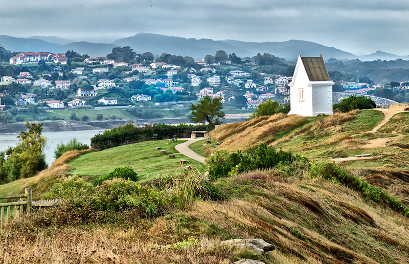 Pequeña capilla blanca sobre una colina cubierta de hierba con vistas a un río, montañas y un pueblo