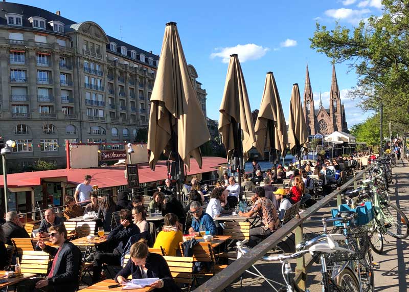 Mucha gente sentada en el Quai des Pecheurs disfrutando de una copa bajo el sol en Estrasburgo