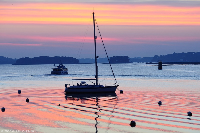 Un velero en una bahía tranquila bajo un cielo con capas de color rosa y morado al atardecer