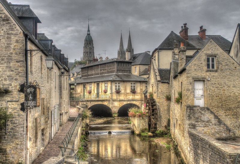 Ciudad de Bayeux en Normandía, las agujas de su catedral asoman sobre casas antiguas, una rueda de molino gira sobre un arroyo