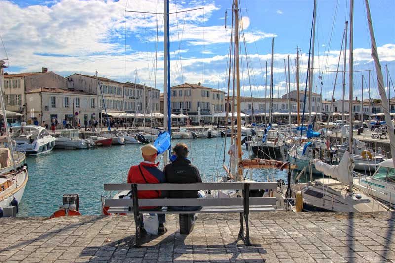 Un hombre y una mujer se sientan en un banco disfrutando de las vistas del puerto de Saint Martin, Ile de Re