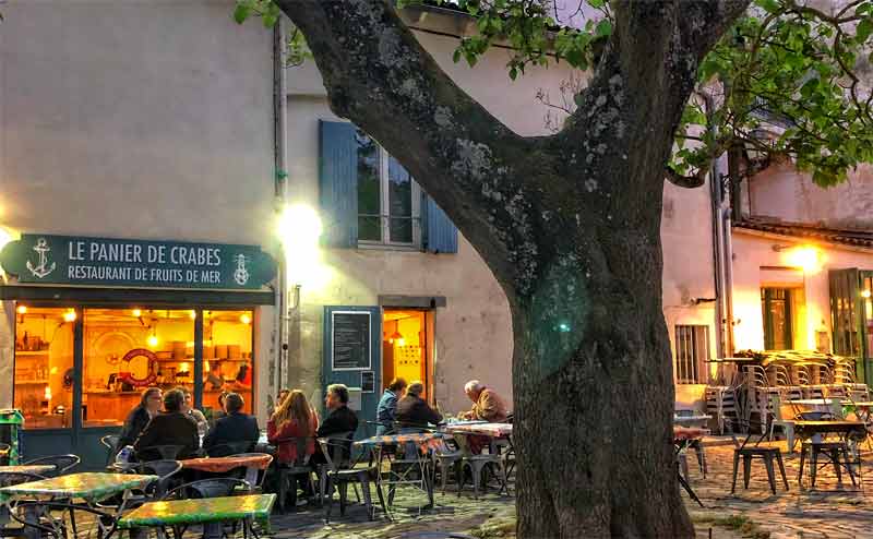 Pequeña plaza con un árbol frondoso y un pequeño restaurante con gente comiendo en mesas en una terraza en La Rochelle