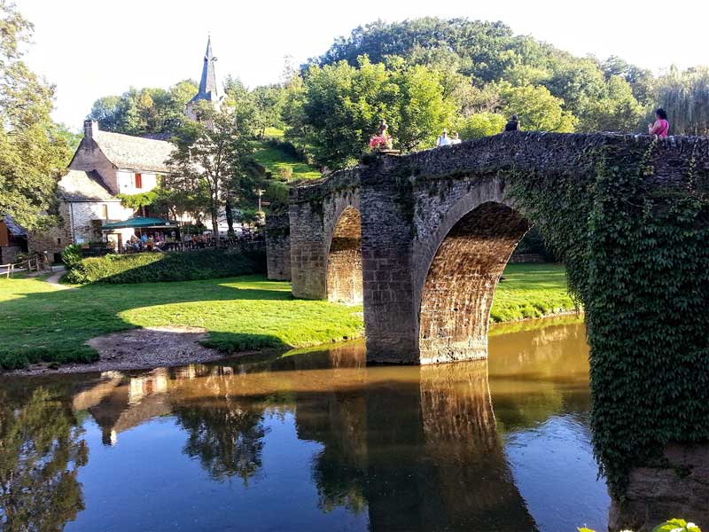 Antiguo puente de piedra arqueado sobre un río que conduce a una ciudad con edificios antiguos y una iglesia, Belcastel
