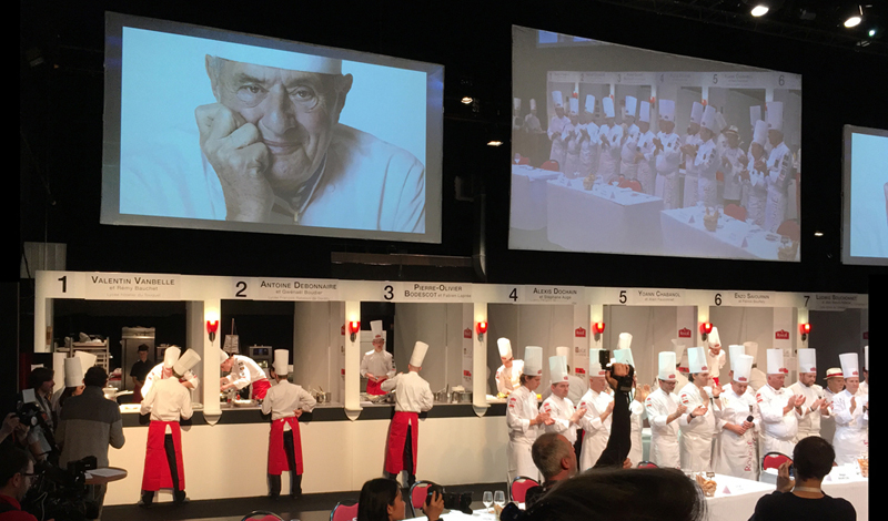 En un amplio escenario, jóvenes chefs con gorros blancos cocinan en un concurso en Sarlat