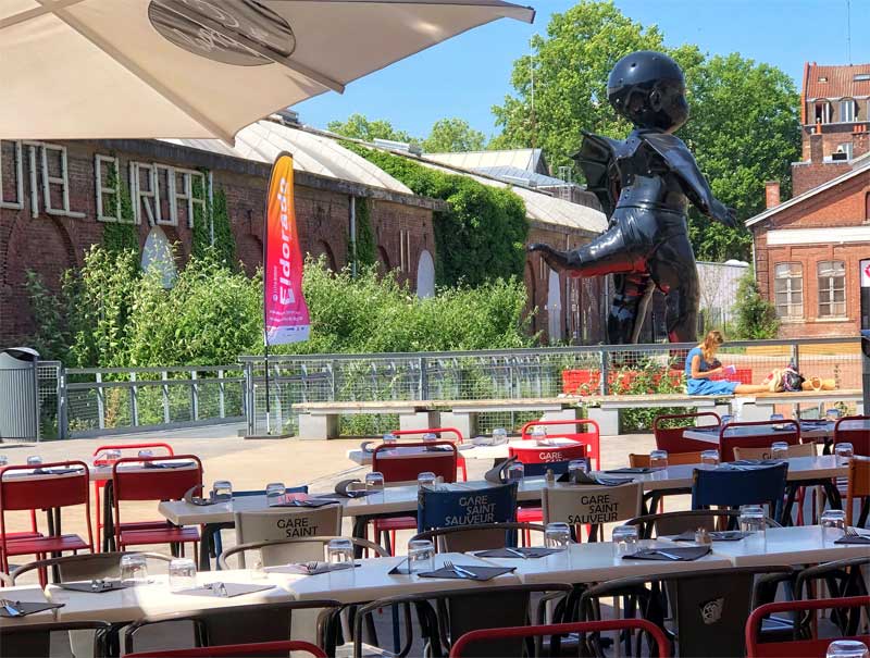Mesas y sillas en una terraza con vistas a una escultura de un bebé con cola gigante en Lille